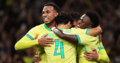 Grupo de jogadores da seleção brasileira, usando uniforme amarelo fluorescente, se abraça em comemoração dentro de campo, sorrindo e celebrando após um gol.