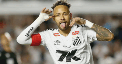 Jogador do Santos comemora gol fazendo gesto com as mãos ao lado do rosto e mostrando a língua; ele veste uniforme branco com braçadeira vermelha e aparece em primeiro plano, com o estádio desfocado ao fundo.