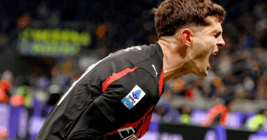 Jogador do Milan, vestindo uniforme preto com detalhes vermelhos, comemora gritando com intensidade, inclinado para frente, com as veias do pescoço saltadas. O fundo mostra arquibancadas desfocadas e luzes do estádio.