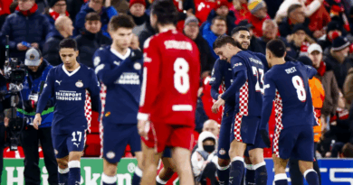 Jogadores do PSV comemoram em campo após marcar gol contra o Liverpool em Anfield, enquanto atletas dos Reds aparecem cabisbaixos ao fundo. A torcida inglesa observa a cena nas arquibancadas.