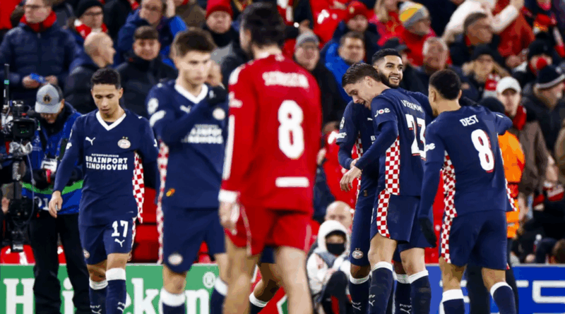 Jogadores do PSV comemoram em campo após marcar gol contra o Liverpool em Anfield, enquanto atletas dos Reds aparecem cabisbaixos ao fundo. A torcida inglesa observa a cena nas arquibancadas.