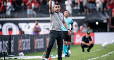 Um técnico do Vitória, à beira do campo, aponta para frente dando instruções durante a partida, com a torcida rubro-negra ao fundo e um árbitro próximo observando.