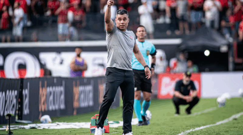 Um técnico do Vitória, à beira do campo, aponta para frente dando instruções durante a partida, com a torcida rubro-negra ao fundo e um árbitro próximo observando.