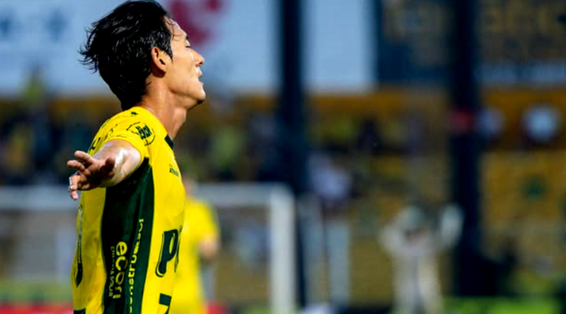 Jogador de futebol com camisa amarela e verde, braços abertos em gesto de comemoração, visto de perfil dentro do estádio, com arquibancadas desfocadas ao fundo durante uma partida noturna.